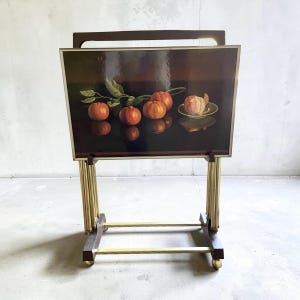 May include: A vintage serving tray table with a dark brown frame and brass-coloured legs. The tray features a still life painting of oranges on a black background. The table has a handle and rolls on small wheels.