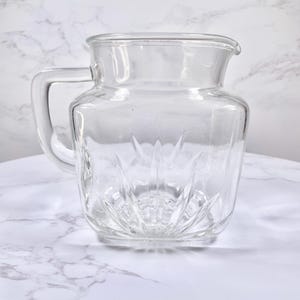 May include: A clear glass jug with a handle and a spout. The jug has a square shape with a decorative pattern on the bottom. The jug is on a white marble surface.