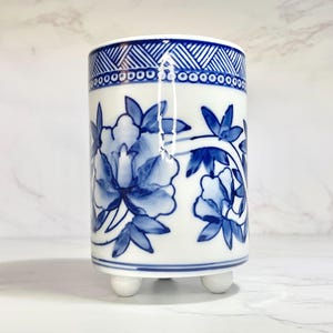 May include: A white ceramic vase with a blue floral design and three small, white feet. The vase features a detailed blue floral pattern and a geometric border at the top. The design is reminiscent of traditional Asian pottery.