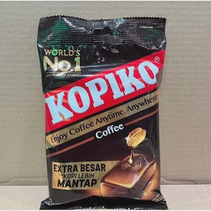 Könnte beinhalten: Eine Packung Kopiko-Kaffeebonbons. Die Verpackung ist hauptsächlich schwarz, rot und braun, mit dem Wort "Kopiko" in großen, hellblauen Buchstaben. Die Verpackung enthält auch den Text "Enjoy Coffee Anytime, Anywhere" und "Extra Besar Kopi Lebih Mantap".