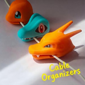 May include: Three colorful cable organizers shaped like creature heads in orange, teal, and orange, each featuring a central slot for a white charging cable. The text Cable Organizers is printed in yellow at the bottom right of the image.