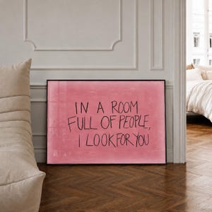 Könnte beinhalten: Ein rechteckiges gerahmtes Wandbild mit einem strukturierten rosa Hintergrund und dem handgeschriebenen schwarzen Text: IN A ROOM FULL OF PEOPLE, I LOOK FOR YOU. Das Kunstwerk ist in einem dünnen schwarzen Rahmen präsentiert.