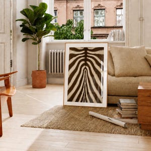 Può includere: Un'opera d'arte murale rettangolare incorniciata con un motivo a strisce di zebra ad alto contrasto in marrone scuro e crema. La stampa è esposta in una cornice di legno chiaro, appoggiata su un tappeto testurizzato in una stanza con pianta.