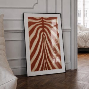 May include: A rectangular wall art print featuring a repeating pattern of rust-coloured and cream-coloured zebra stripes. The print is encased in a thin black frame and displays a high-gloss finish that reflects light across the surface of the artwork.