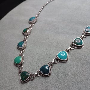 May include: Silver necklace featuring turquoise-colored, triangular gemstones. The stones are set in a silver bezel with a beaded edge. The necklace has a delicate chain and a variety of turquoise shades, from light to dark.