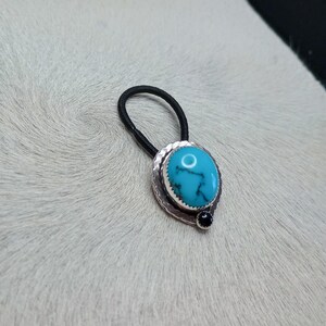 May include: Oval turquoise stone hair tie with a silver-colored bezel and a small black stone accent. The hair tie has a black elastic band. The turquoise stone is a bright, vibrant blue-green color.