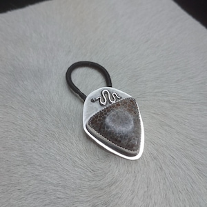 May include: A silver-toned hair tie featuring a patterned stone pendant. The pendant has a mix of brown, grey, and white tones, with a silver snake design at the top. The hair tie is black.