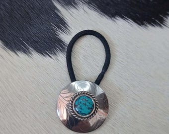 May include: Silver-toned hair tie with a turquoise stone center. The round pendant features a detailed floral design and a twisted rope border around the stone. The hair tie has a black elastic band.