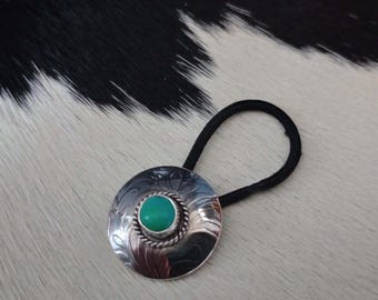May include: Silver-toned hair tie with a circular, engraved design. The center features a round, turquoise-colored stone. The hair tie has a black elastic band. The design is reminiscent of Southwestern or Western jewelry.