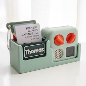 May include: A mint green toy radio and book holder. The holder has a compartment with a book titled "The Very Hungry Caterpillar & Other Stories" and the name "Thomas". The radio has orange knobs and a speaker.