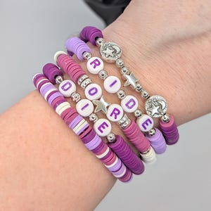 May include: Six handmade bracelets in shades of purple, pink, and white. The bracelets are made of flat, round beads and feature silver accents and letter beads spelling out "ROIDE". Some bracelets have star charms.
