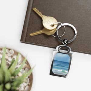 Può includere: Un portachiavi argentato con un ciondolo rettangolare raffigurante un dipinto di paesaggio marino con onde blu dell'oceano sotto un cielo blu. Due chiavi d'oro sono attaccate, appoggiate su una superficie marrone.