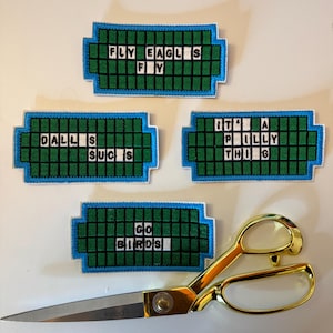 May include: Four embroidered patches with text, including "FLY EAGLES FLY", "DALLAS SUCKS", "IT'S A PHILLY THING", and "GO BIRDS". A pair of gold scissors is positioned below the patches.