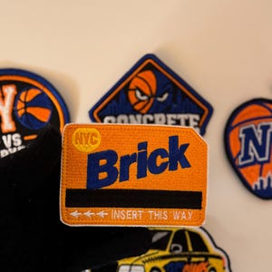 May include: A rectangular embroidered patch with an orange background, featuring the text "NYC" in a yellow circle, the word "Brick" in large blue letters, a black horizontal stripe, and the white text "INSERT THIS WAY" with three arrows.