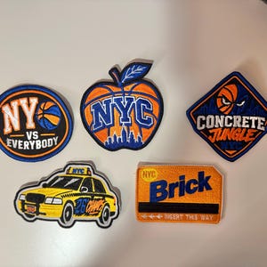 May include: Five embroidered patches featuring New York City themes. One circular patch reads NY VS EVERYBODY with a basketball. An apple-shaped patch shows a basketball and NYC skyline. A diamond patch says CONCRETE JUNGLE NYC. A yellow taxi patch and a rectangular patch reading Brick with NYC and INSERT THIS WAY.