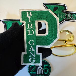 May include: Green and white embroidered patches with the text "BIRD GANG" and "215 PHILLY". The patches include a large green letter "P" and a green letter "B". Also included are gold-rimmed eyeglasses.