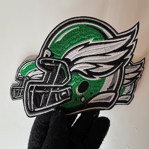 May include: Embroidered patch depicting a green and black American football helmet with white wings. The helmet has a black facemask and is outlined in black and white. Suitable for sports team or fan apparel.