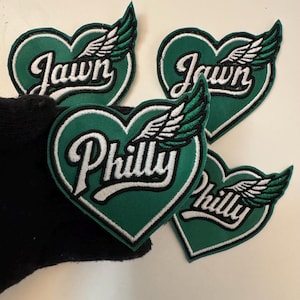 May include: Four heart-shaped patches with green backgrounds and white borders. Each patch features the word "Jawn" or "Philly" in white script, along with a white and green winged design. These patches are likely for clothing or accessories.