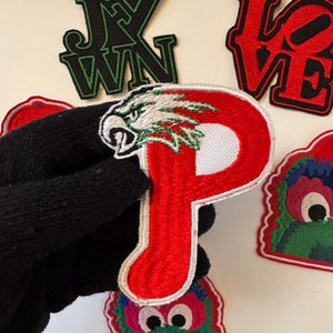 May include: A collection of embroidered iron-on patches. The patches feature a red letter "P" with a white eagle head, a red "LOVE" patch, a black and green patch with a monster face, and a black patch with green lettering that says "J.X.W.N."