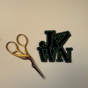 May include: A black and green iron-on patch with the text "JXN" in a bold, blocky font. A pair of gold scissors are also in the image.