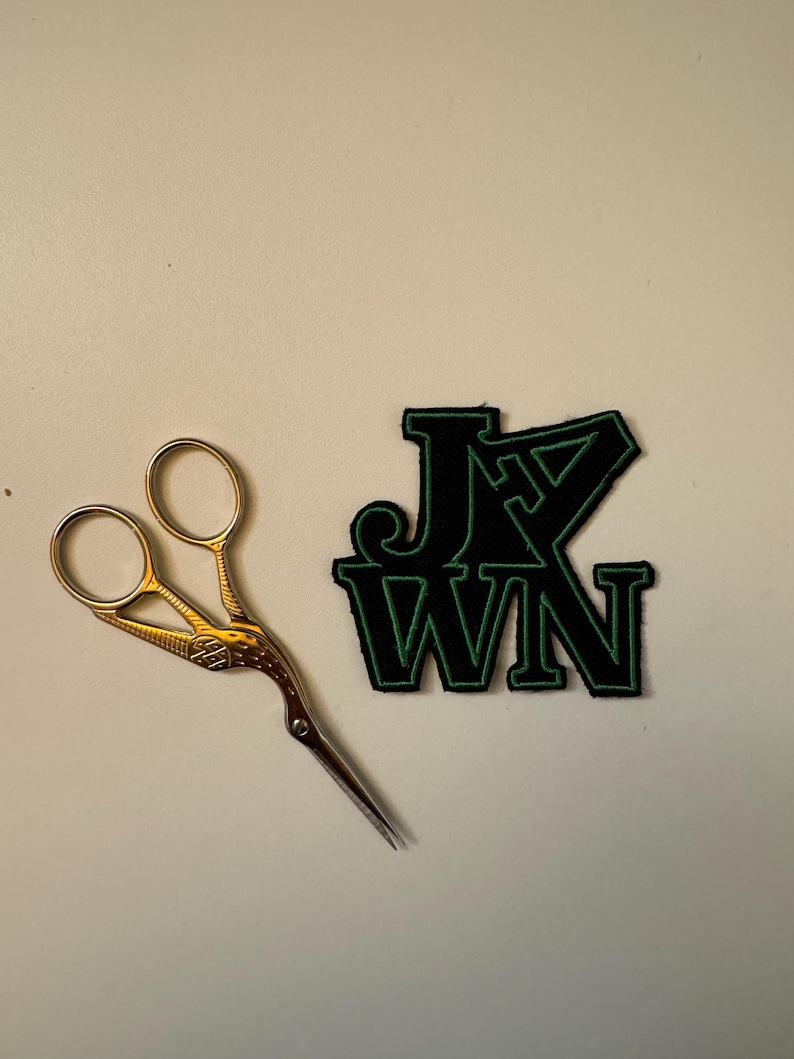 May include: A black and green iron-on patch with the text "JXN" in a bold, blocky font. A pair of gold scissors are also in the image.