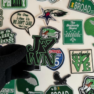 May include: A collection of green and black embroidered patches with various designs, including a football helmet, a star, an eagle, and a street sign that reads "99 N BROAD ST".