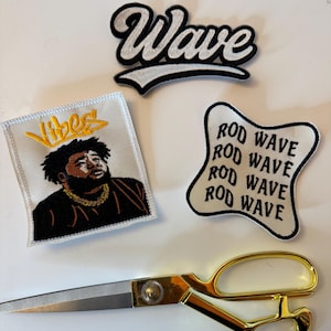 May include: An assortment of embroidered patches with the words "Wave" and "Vibes", and a portrait of a person. A pair of gold-handled scissors is also present. The patches are in black, white, and gold.