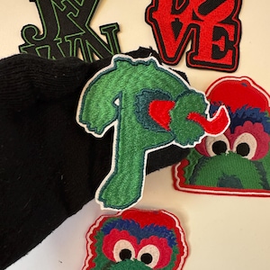 May include: A collection of colorful iron-on patches featuring a green shirt, a red heart, a red and green monster with big eyes, and the word "LOVE" in red with black outlines.