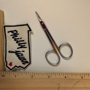 May include: A pair of small, silver scissors with a red accent, next to a cream-colored patch with the words "Philly Jawn" in black script and a red heart. A wooden ruler with inch markings is in the foreground.