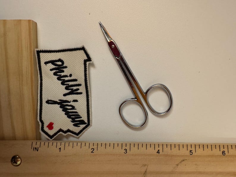 May include: A pair of small, silver scissors with a red accent, next to a cream-colored patch with the words "Philly Jawn" in black script and a red heart. A wooden ruler with inch markings is in the foreground.