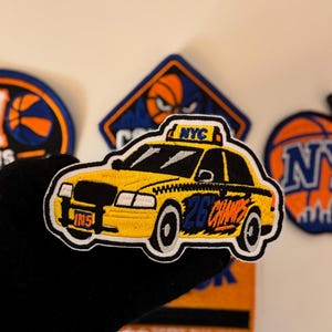 May include: An embroidered patch shaped like a yellow New York City taxi cab. The taxi features a roof sign reading NYC, a front license plate reading INS, and the text 26 CHAMPS in blue and orange on the side. The patch has a black border.