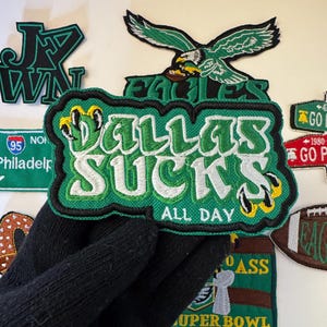 May include: A collection of embroidered patches with sports-related themes. The central patch reads "DALLAS SUCKS ALL DAY" in green and white, with a black border. Other patches include an eagle, football, and text like "SUPER BOWL LIX CHAMPS".