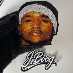 Peut inclure: Écusson brodé représentant un portrait d'une personne avec une casquette noire et une garniture argentée. L'écusson a une bordure noire. En dessous, un écusson séparé avec le nom "J Boog" en écriture blanche avec un contour noir.