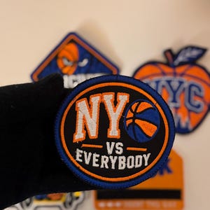 Puede incluir: Un parche bordado circular con fondo negro y borde naranja. El texto NY VS EVERYBODY aparece en letras blancas y naranjas, acompañado de un gráfico de un balón de baloncesto en colores azul y naranja.