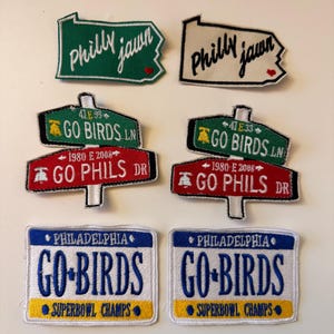May include: A collection of embroidered patches featuring Philadelphia sports team themes. Includes patches with "Philly Jawn", "Go Birds", "Go Phils", and "Superbowl Champs" text. The patches are in green, red, blue, and white.