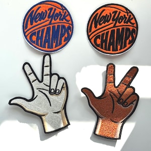 May include: Four embroidered patches including two circular basketball-themed patches with the text New York CHAMPS in orange and blue, and two hand-shaped patches displaying a three-finger gesture in light and dark skin tones with black outlines.