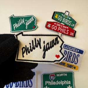 May include: A collection of embroidered patches with Philadelphia-themed designs. The patches feature phrases like "Philly jawn," "Go Birds," and "Go Phils," along with a license plate design and a road sign. The patches are in various shapes and colors.