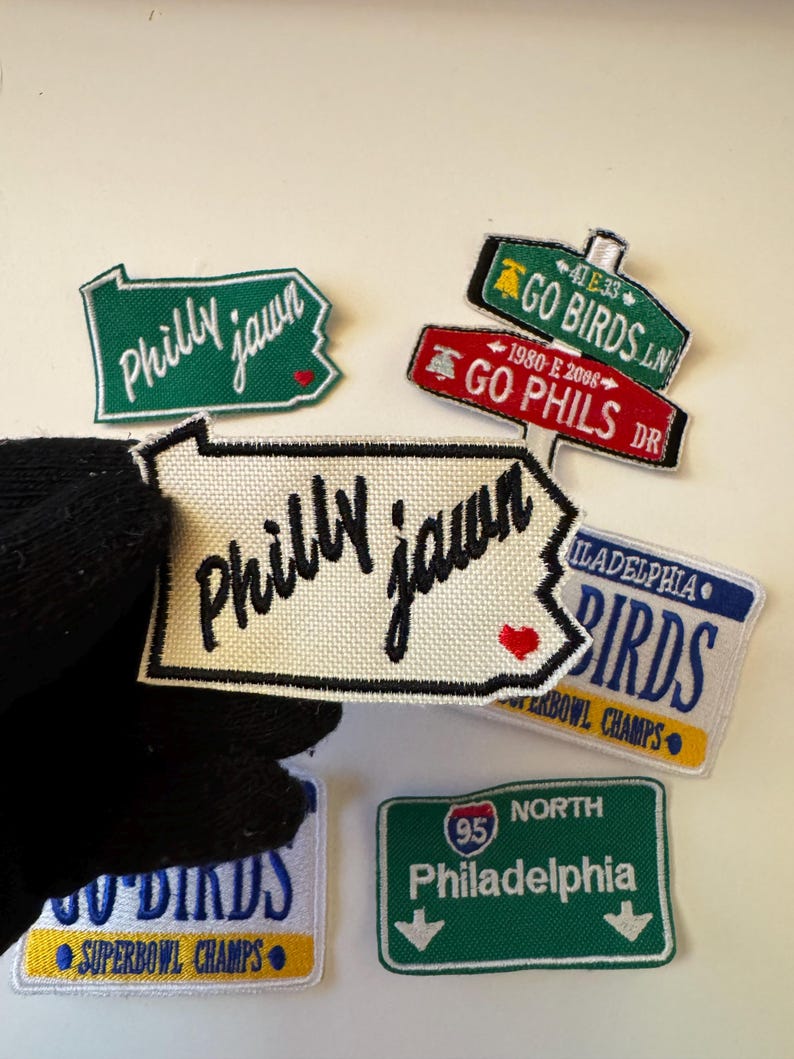 May include: A collection of embroidered patches with Philadelphia-themed designs. The patches feature phrases like "Philly jawn," "Go Birds," and "Go Phils," along with a license plate design and a road sign. The patches are in various shapes and colors.