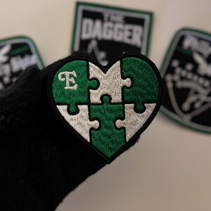 May include: A heart-shaped embroidered patch featuring a four-piece jigsaw puzzle design in alternating dark green and silver-white colours. The top-left puzzle piece includes a white embroidered letter E. The patch has a thick black border.