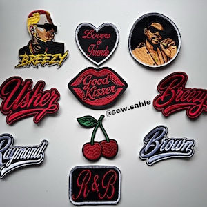 May include: A collection of embroidered patches featuring various designs including portraits of a man, red lips with the text Good Kisser, a cherry motif, and script text patches reading Breezy, Usher, Raymond, Brown, Lovers & Friends, and R&B.