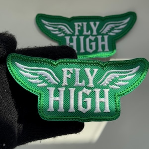 Puede incluir: Dos parches bordados verdes con alas blancas a los lados del texto FLY HIGH en una fuente en mayúsculas. Los parches tienen una superficie de tela texturizada y un borde cosido, con un parche en primer plano y otro situado detrás.
