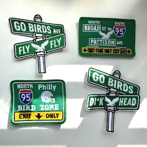 May include: Four embroidered patches featuring Philadelphia sports themes. Two patches are shaped like street signs with text reading GO BIRDS AVE and FLY FLY or DI*K HEAD. Two rectangular patches resemble highway signs with text like BIRD ZONE.