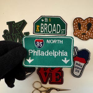 May include: Assortment of iron-on patches featuring various designs, including a pretzel, a Philly water ice cup, a "Love" patch, a "99 N Broad St" street sign, a "Philadelphia" highway sign, and a "JXV" patch. The patches are arranged on a white surface with a pair of gold scissors.