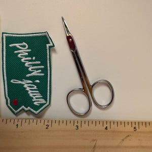 May include: A green and white embroidered patch with the text "Philly Jawn" and a small red heart. Next to it is a pair of small, silver scissors with a red accent. A wooden ruler is in the foreground.