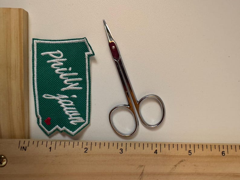 May include: A green and white embroidered patch with the text "Philly Jawn" and a small red heart. Next to it is a pair of small, silver scissors with a red accent. A wooden ruler is in the foreground.