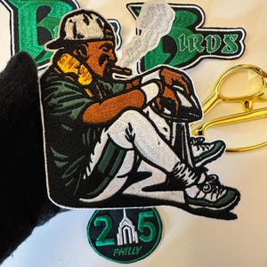 May include: Embroidered patch featuring a seated figure smoking a cigar, wearing a baseball cap and jersey. The patch is in green, white, brown, and black. Additional patches include the word "BIRDS" and a "215 PHILLY" patch.