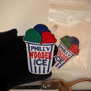 May include: Two embroidered patches shaped like a cup of water ice. The design features a white cup with blue stripes and the text PHILLY WOODER ICE in blue and red. The top shows scoops of green, blue, and red ice with a small white spoon.