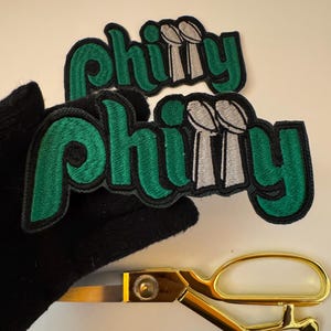May include: Two embroidered "Philly" patches in green and black, featuring silver trophy designs. Gold scissors are in the foreground.