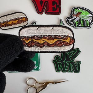 May include: A collection of embroidered iron-on patches featuring a hot dog, the words "LOVE", "FLYIN", and "J.K. WIN", and a pair of gold scissors.