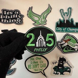 Peut inclure: Une collection de patchs brodés verts et noirs représentant le logo des Philadelphia Eagles, les toits de la ville et diverses phrases comme "It's a Philly thing", "25 Philly", "No One Likes Us We Don't Care", et "Fly".